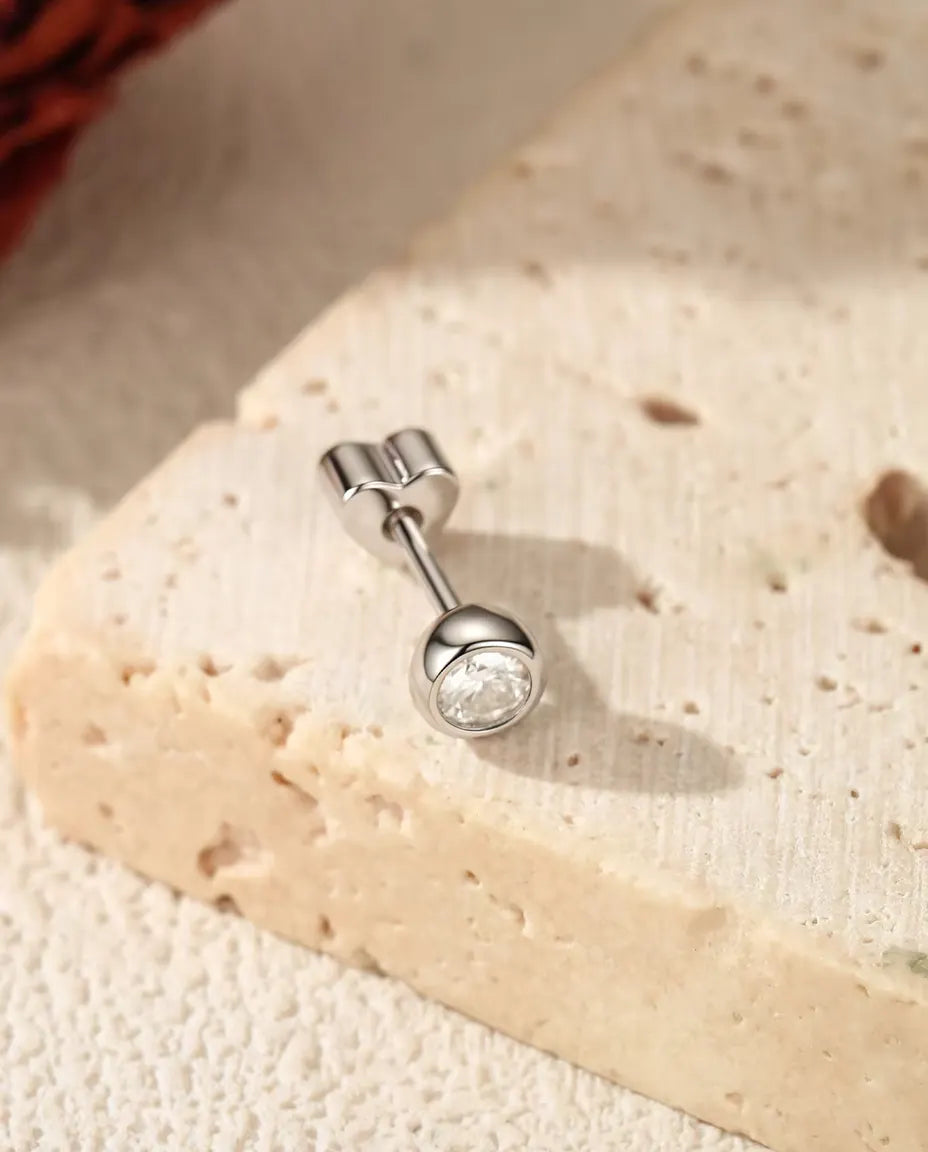 Orecchini con Moissanite Tonda da 3 mm in Argento 925 - su una superficie chiara ruvida accanto a una lastra di travertino