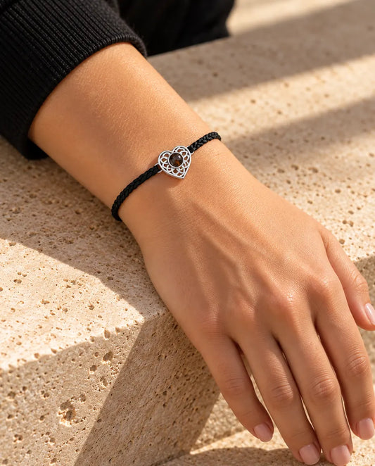 Donna che indossa al polso il bracciale con cordino intrecciato sottile nero e cuore argentato, alla luce calda