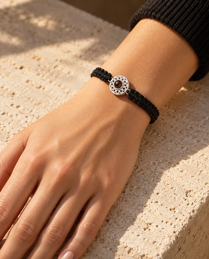 Donna che indossa al polso il bracciale con cordino intrecciato nero e charm tondo argentato, alla luce calda