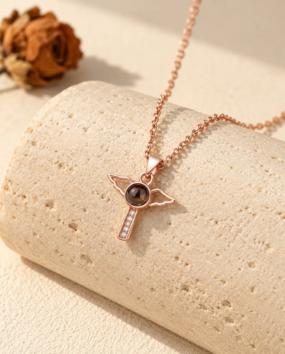 Collana color oro rosa con croce e ali d'angelo appesa davanti a un blocco di travertino