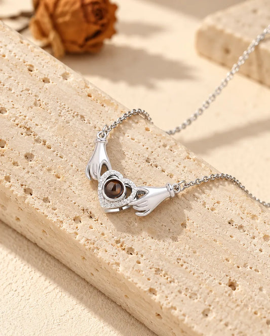 Collana con foto dentro argentata con due piccole mani che tengono un cuore a pavé, appoggiata su un blocco di travertino