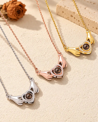 Collane con due mani che tengono un cuore, in versione argentata, color oro rosa e dorata, una accanto all'altra