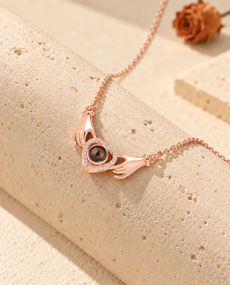 Collana con foto dentro color oro rosa con due mani che tengono un cuore, adagiata su un blocco di travertino
