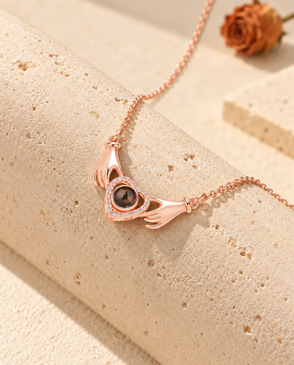Collana con foto dentro color oro rosa con due mani che tengono un cuore, adagiata su un blocco di travertino
