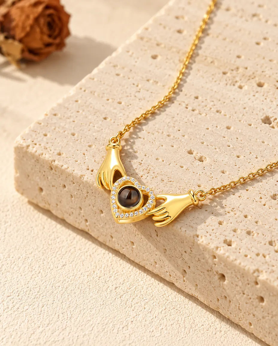 Collana dorata con foto dentro e due mani che stringono un cuore di zirconi, su una lastra di travertino