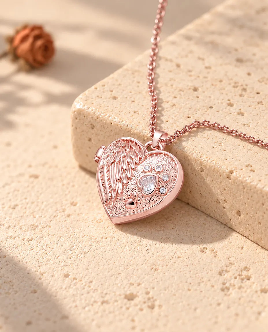 Collana portafoto color oro rosa a cuore con ala d'angelo e zampa sul travertino, nella calda luce del sole