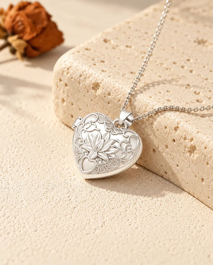 Collana portafoto argentata a cuore con fiore di loto e volute in rilievo, appoggiata su un blocco di travertino