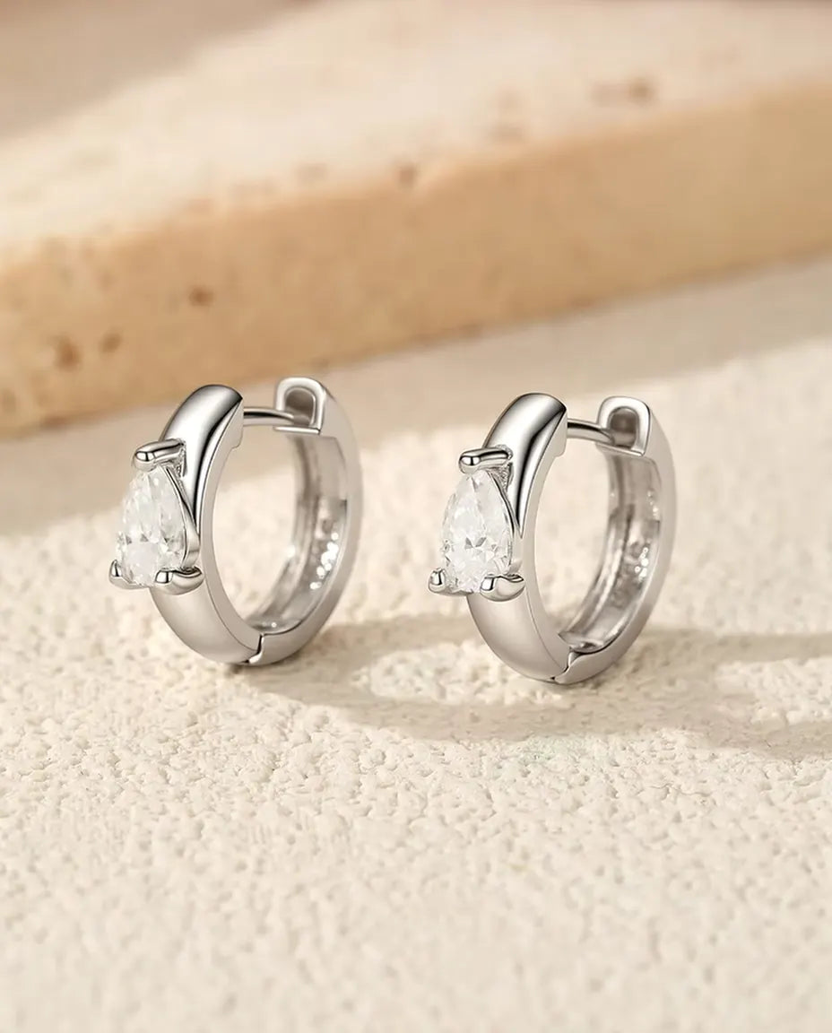 Orecchini a Cerchio Huggie con Moissanite a Goccia in Argento 925 - la coppia in piedi, uno accanto all’altro, su una superficie chiara ruvida