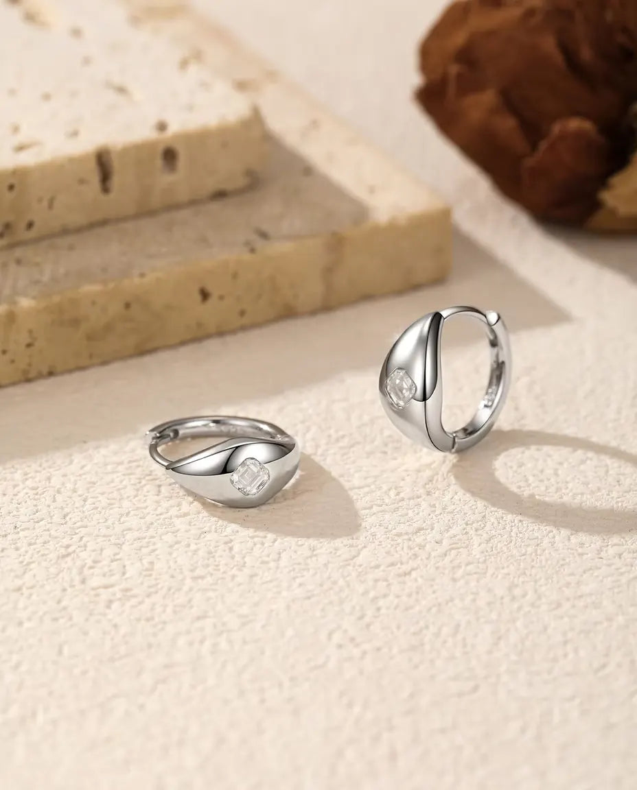 Orecchini a Cerchio Huggie Moissanite Asscher in Argento 925 - la coppia su una superficie chiara ruvida, uno disteso e uno in piedi