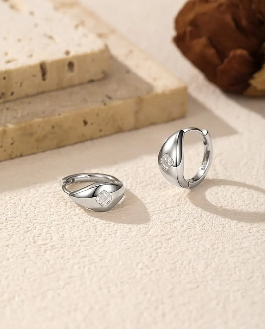 Orecchini a Cerchio Huggie Moissanite Asscher in Argento 925 - la coppia su una superficie chiara ruvida, uno disteso e uno in piedi