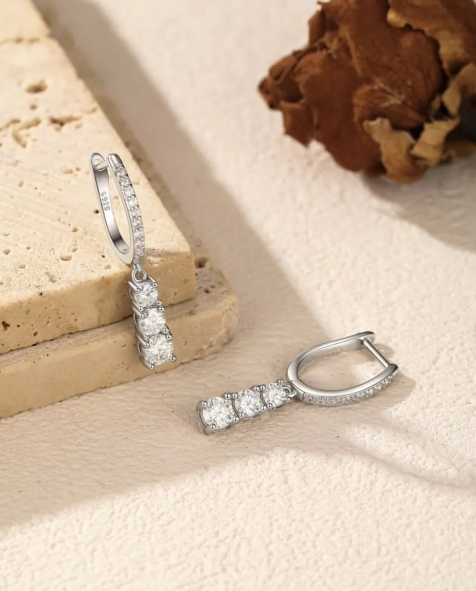 Orecchini a Cerchio Pendenti Moissanite a Tre Pietre in Argento 925 - un orecchino appeso a una lastra di travertino, l’altro disteso accanto