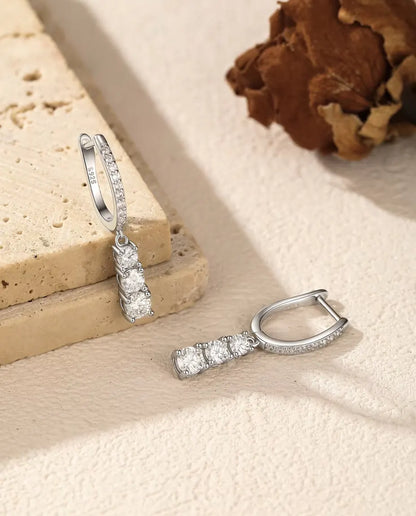 Orecchini a Cerchio Pendenti Moissanite a Tre Pietre in Argento 925 - un orecchino appeso a una lastra di travertino, l’altro disteso accanto