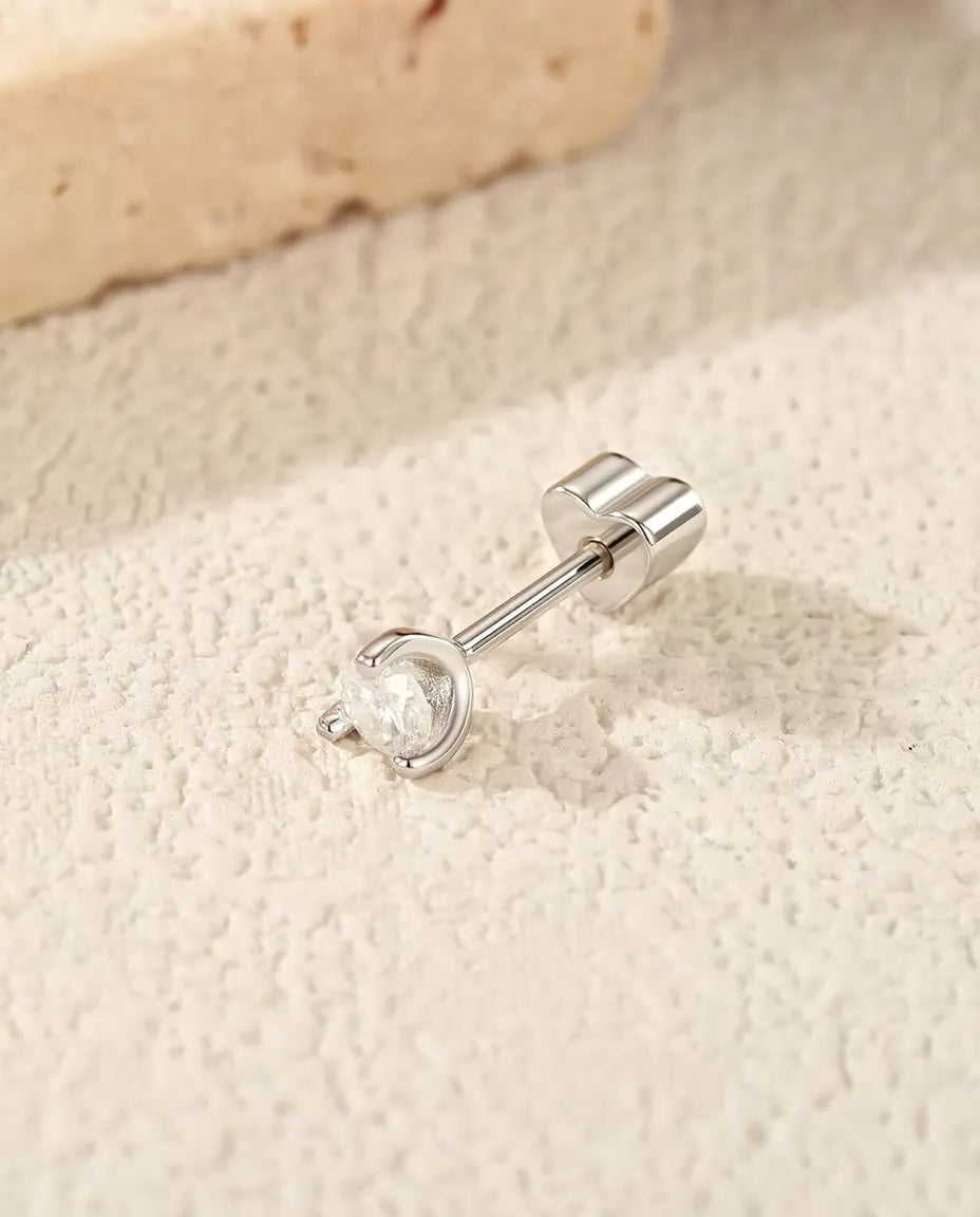 Orecchini con Moissanite Tonda e Arco Laterale in Argento 925 - appoggiato di lato su una superficie chiara ruvida, chiusura a forma di cuore sul perno